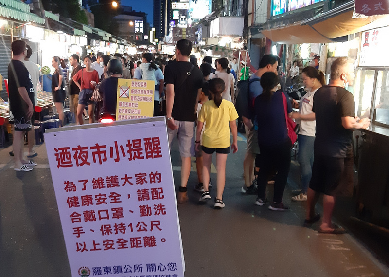 中央流行疫情指揮中心宣布8大場所戴口罩後的首個週末，宜蘭縣羅東鎮公所在夜市出入口放上告示牌，提醒民眾戴起口罩並維持社交距離。