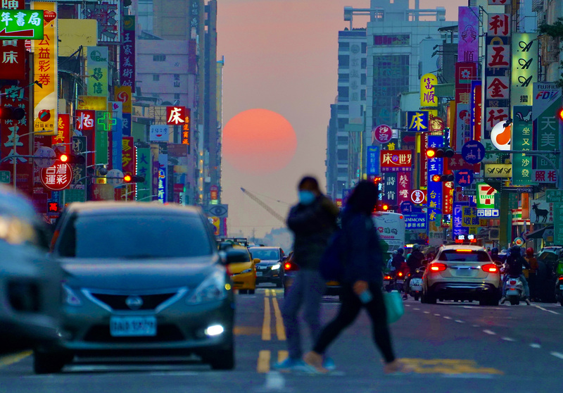 高雄市青年路「懸日」美景28日傍晚再度現身，橘澄澄的太陽緩緩落下，以車水馬龍的街道與五光十色的招牌為襯，格外吸睛，也吸引許多攝影愛好者前往搶拍。 (圖：中央社)