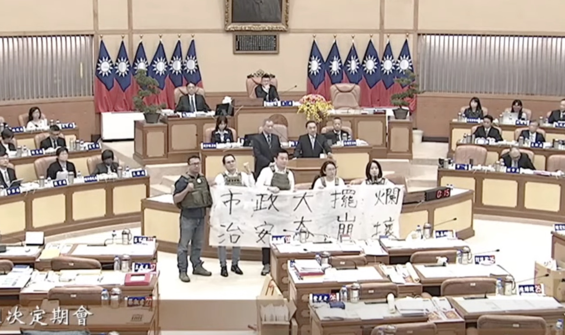 新北市議會民進黨團議員11日身穿防彈背心，在市政總質詢中拉白布條要求市長侯友宜進行「治安專案報告」。(圖片來源／新北市議會)
