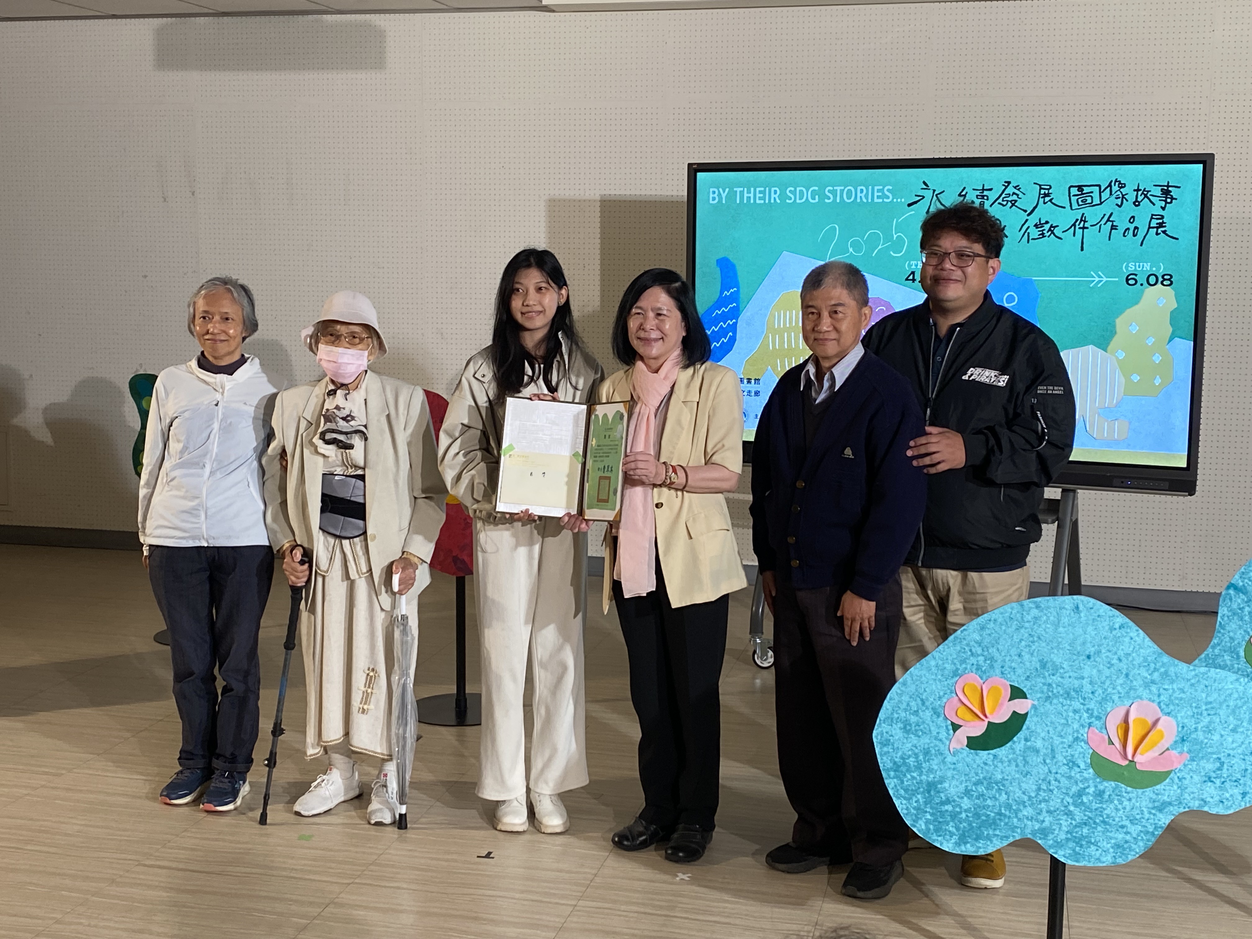 國立台灣圖書館25日舉辦「永續發展圖像故事與徵件作品展」開幕活動，館長曹翠英頒發獎狀及新台幣6萬元獎金給「永續發展圖像徵件首獎得主周品劭，周品劭的親友特別到場共享喜悅並上台合影留念。(陳念宜 攝)